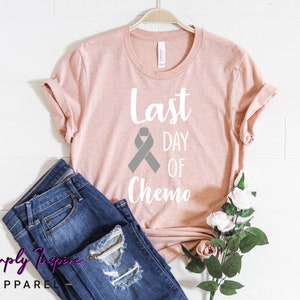 May include: A pink t-shirt with the text "Last Day of Chemo" and a grey ribbon. The shirt is on a white background with a pair of blue jeans and a bouquet of white roses.