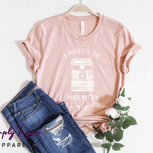 May include: A pink t-shirt with the text "A photo is the pause button of life" and a white outline of an instant camera.