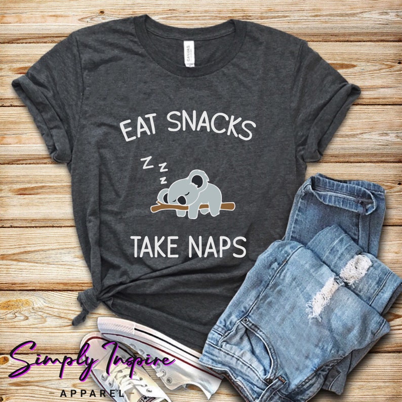 May include: A dark gray t-shirt with the text "EAT SNACKS" and "TAKE NAPS" in white. A cartoon koala is sleeping on a branch between the two lines of text. The text "Z Z Z" is above the koala.