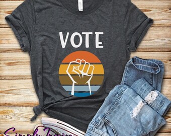 May include: A dark grey t-shirt with the word "VOTE" in white text and a raised fist graphic with a retro sunset design.