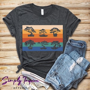 May include: Dark gray t-shirt featuring a graphic of six bonsai trees in silhouette against a retro striped background of orange, tan, red, and blue. The shirt is paired with blue jeans and white sneakers.