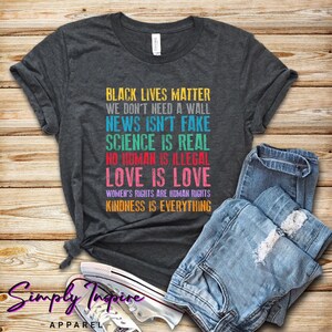 Puede incluir: Una camiseta gris oscuro con un estampado gráfico de arcoíris que dice "Black Lives Matter, We Don't Need a Wall, News Isn't Fake, Science Is Real, No Human Is Illegal, Love Is Love, Women's Rights Are Human Rights, Kindness Is Everything".