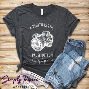 May include: A dark gray t-shirt with a white graphic of a camera and the text "A photo is the pause button of life".