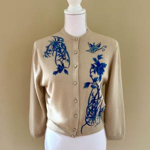 May include: A tan cardigan with three-quarter sleeves and a round neckline. The sweater features blue floral and butterfly embroidery. The cardigan is buttoned up and displayed on a mannequin.