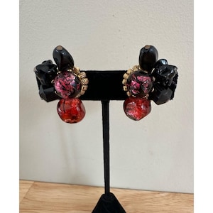May include: A pair of vintage clip-on earrings. Each earring features a cluster of black, faceted beads and a red, glass-like bead. The center bead has a pink and red speckled design, surrounded by gold-toned metal. The earrings are displayed on a black stand.