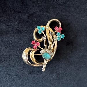 May include: A gold-toned brooch with a floral design. The brooch features a cluster of small, colorful gemstones in shades of blue, pink, and clear, arranged to resemble flowers. The brooch has a curved, elegant shape.