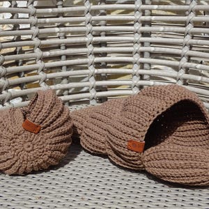 Op de afbeelding: Twee bruine gebreide items liggen op een rieten stoel. Eén is een ronde, gesloten vorm met een leren label. De andere is een grotere, open, schelpachtige vorm, eveneens met een leren label. Beide zijn gemaakt van dik, getextureerd garen.