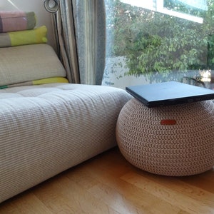 Pu&ograve; includere: Un pouf in crochet beige con un computer portatile sopra. Il pouf &egrave; seduto su un pavimento di legno di fronte a una finestra con vista su alberi verdi.