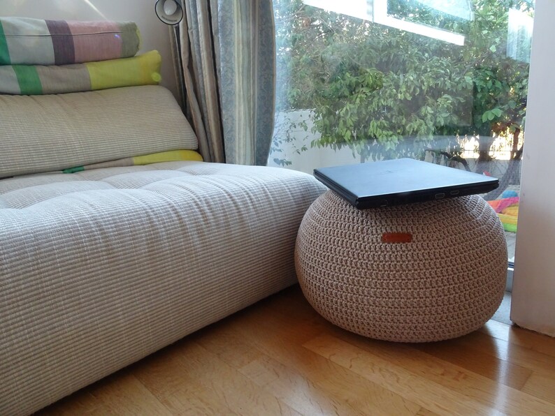 Pu&ograve; includere: Un pouf in crochet beige con un computer portatile sopra. Il pouf &egrave; seduto su un pavimento di legno di fronte a una finestra con vista su alberi verdi.