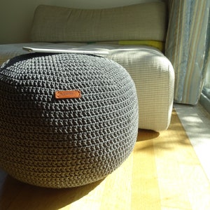 Pu&ograve; includere: Un pouf rotondo in crochet grigio con un'etichetta in pelle marrone che dice "Handmade".