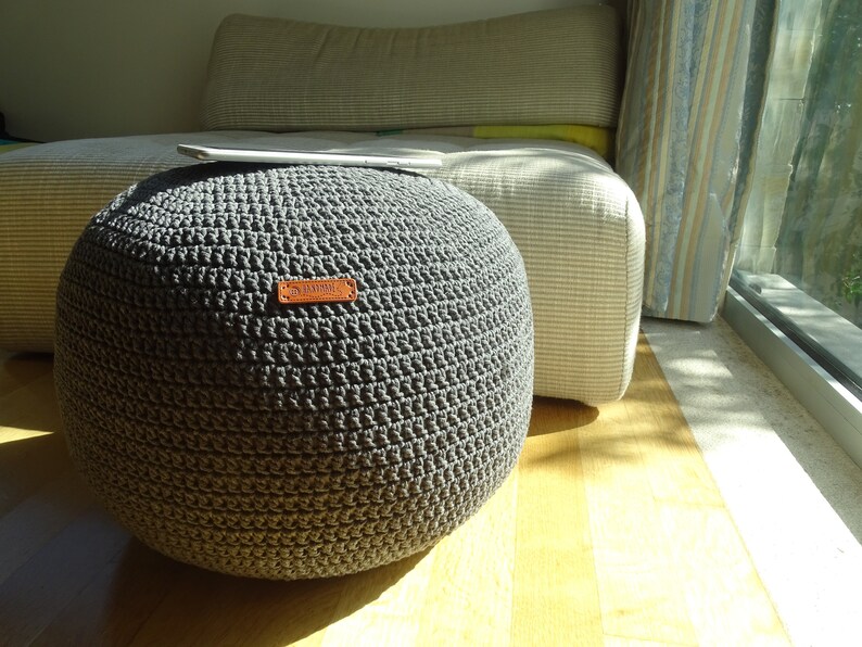 Pu&ograve; includere: Un pouf rotondo in crochet grigio con un'etichetta in pelle marrone che dice "Handmade".