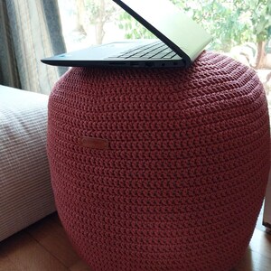 Op de afbeelding: Een grote, ronde, roodbruine gehaakte poef met een laptop erop. De poef is gemaakt van dik garen en heeft een gestructureerd oppervlak. De laptop is zwart en open. De poef heeft een diameter van ongeveer 61 cm.