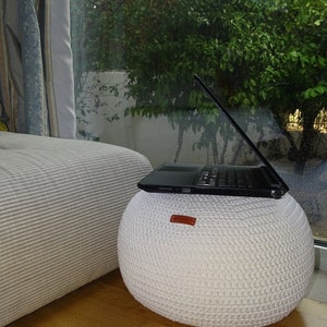 Pu&ograve; includere: Un pouf bianco lavorato a maglia con una maniglia in pelle marrone, un computer portatile nero &egrave; appoggiato sul pouf.