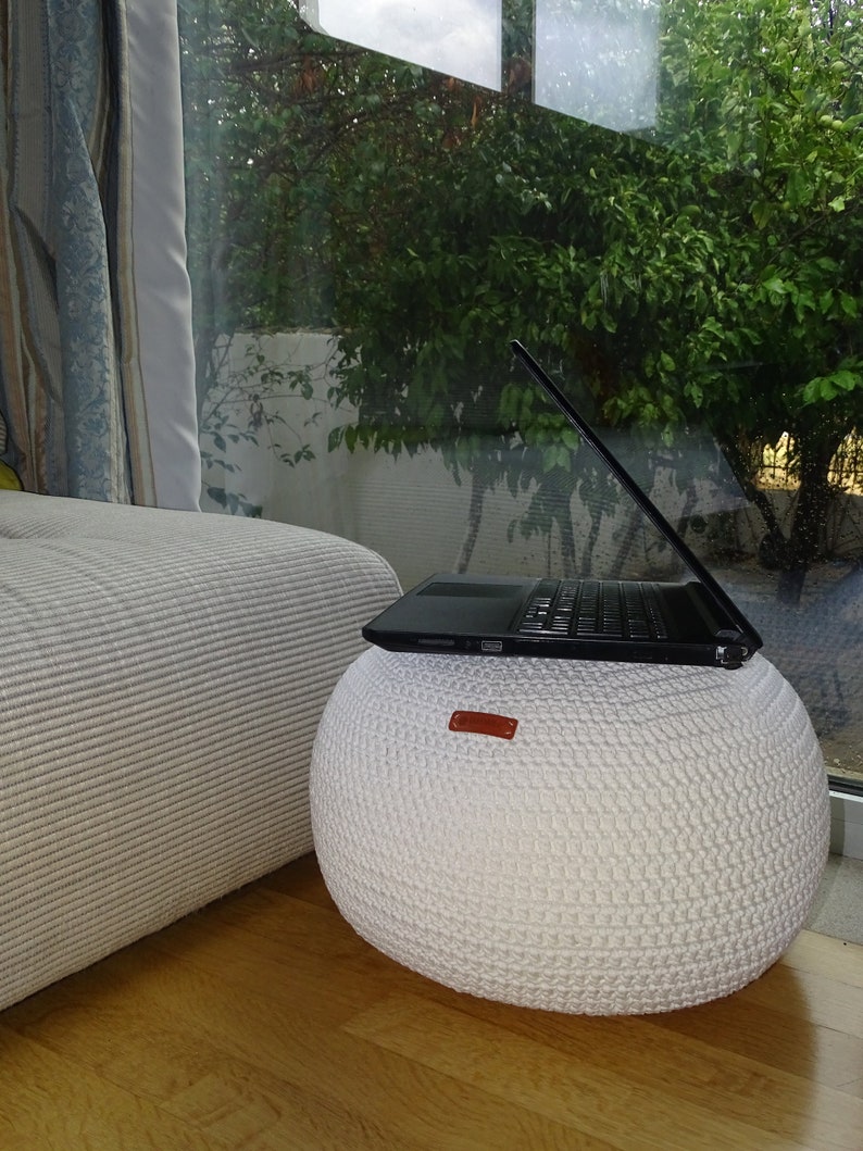 Pu&ograve; includere: Un pouf bianco lavorato a maglia con una maniglia in pelle marrone, un computer portatile nero &egrave; appoggiato sul pouf.