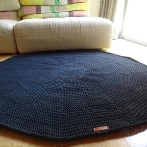 May include: A dark blue, round, crocheted rug with a geometric pattern. The rug is placed on a wooden floor, with a couch and pillows in the background. A small, rectangular tag is sewn on the rug.