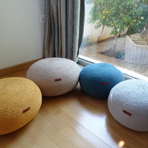 May include: Four round crocheted poufs in different colours: mustard yellow, light grey, light grey, and teal blue. The poufs are arranged in a semi-circle on a wooden floor.