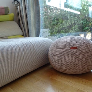 Pu&ograve; includere: Un pouf in crochet color beige chiaro con un'etichetta in pelle. Il pouf &egrave; posizionato su un pavimento in legno davanti a un divano bianco.