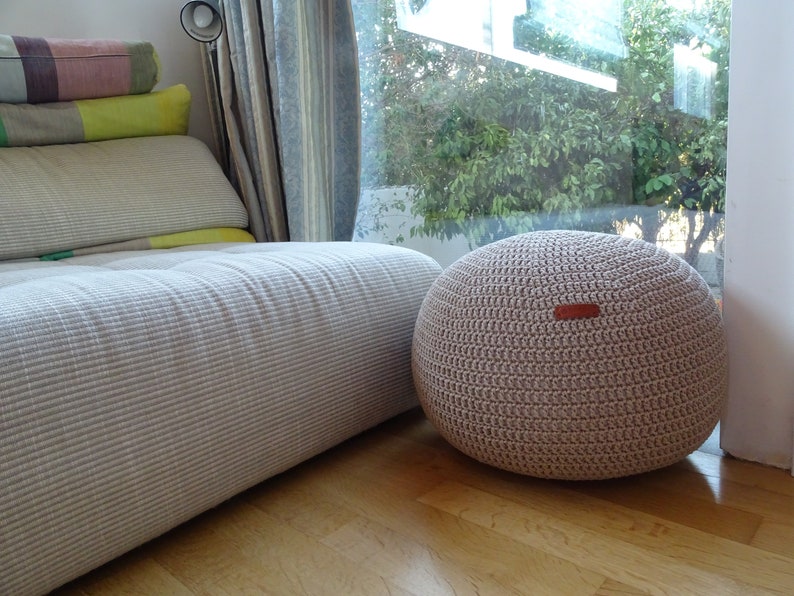 Pu&ograve; includere: Un pouf in crochet color beige chiaro con un'etichetta in pelle. Il pouf &egrave; posizionato su un pavimento in legno davanti a un divano bianco.