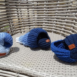 May include: Three handmade crochet items in shades of blue, resembling seashells. The items include a curled hat, a shell-shaped cave, and a small basket. Each piece has a brown leather tag. The items are displayed on a woven chair.