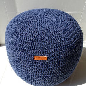 Pu&ograve; includere: Un pouf in crochet blu navy con un'etichetta in pelle marrone che dice "Handmade".