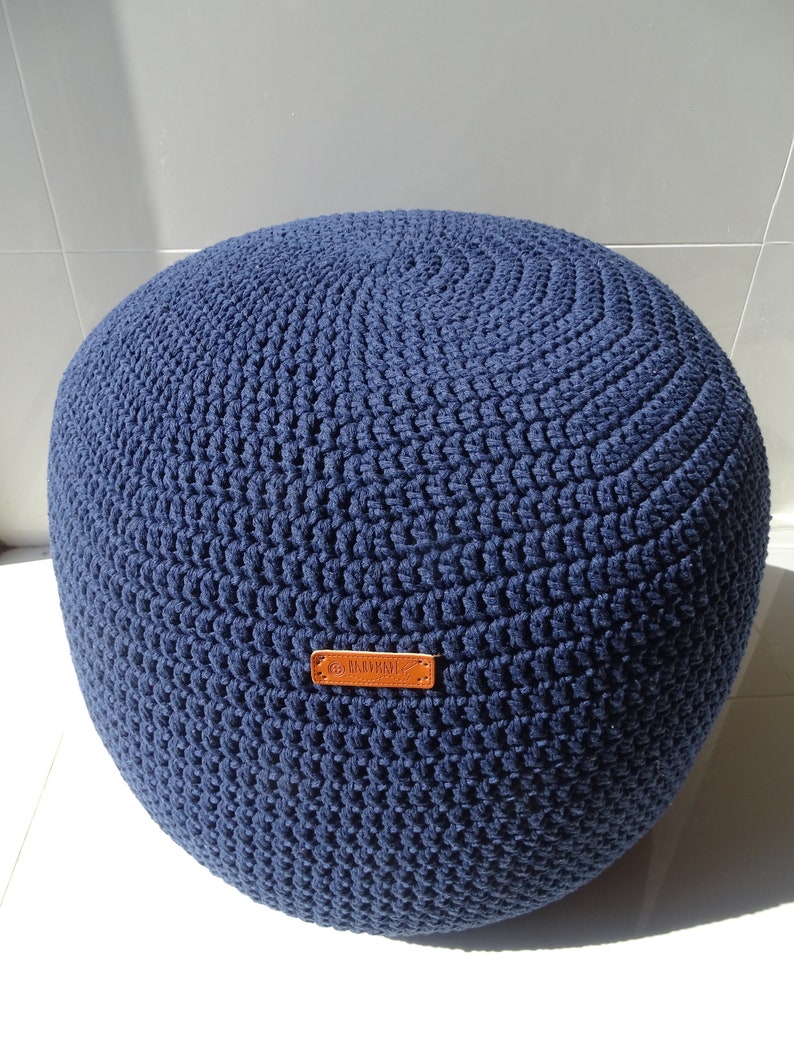 Pu&ograve; includere: Un pouf in crochet blu navy con un'etichetta in pelle marrone che dice "Handmade".