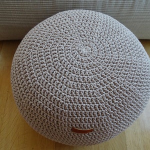 Pu&ograve; includere: Un pouf rotondo in crochet beige con un'etichetta in pelle marrone.