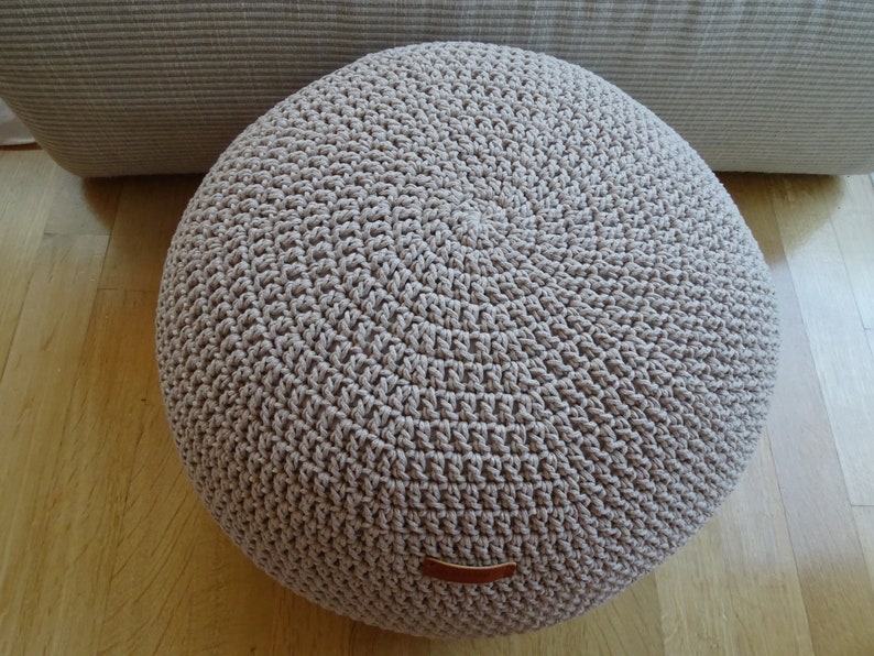 Pu&ograve; includere: Un pouf rotondo in crochet beige con un'etichetta in pelle marrone.