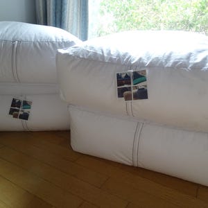 Op de afbeelding: Vier witte vierkante vloerkussens gestapeld in twee paren. Elk kussen heeft een centrale verticale naad met donkere bies en een klein vierkant label aan de voorzijde, met een raster van vier kleine afbeeldingen van diverse meubelstukken.