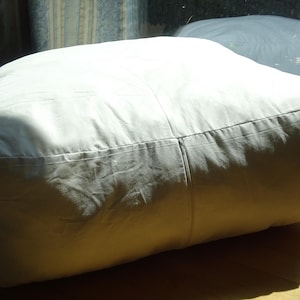 Puede incluir: Un puf grande y cuadrado de color blanco roto. El puf está hecho de una tela suave y tiene un cierre de cremallera. El puf está sobre un suelo de madera, con la luz del sol proyectando sombras sobre su superficie.