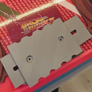 Pode incluir: Uma prancha voadora com o tema De Volta para o Futuro com componentes de plástico cinza e o logotipo do filme. A prancha tem um design rosa e vermelho com uma régua na borda. Parafusos são visíveis.