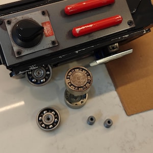 May include: A black and silver toy gun with red buttons and a black knob. The gun is disassembled and the parts are laid out on a white surface. The parts include a silver gear, a silver bearing, and two silver washers.