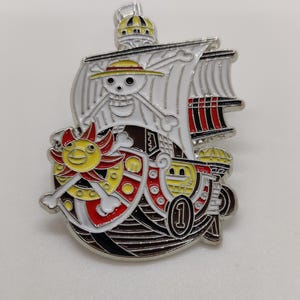 May include: A metal enamel pin shaped like a ship with a white sail featuring a skull and crossbones wearing a yellow straw hat. The ship has a red and yellow sun emblem, a black hull, and a circular number one symbol on the side of the vessel.