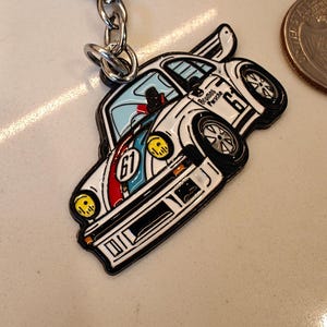 May include: White metal keychain featuring a cartoon depiction of a blue and red race car with the text "Blumos Porsche 67" on the side.