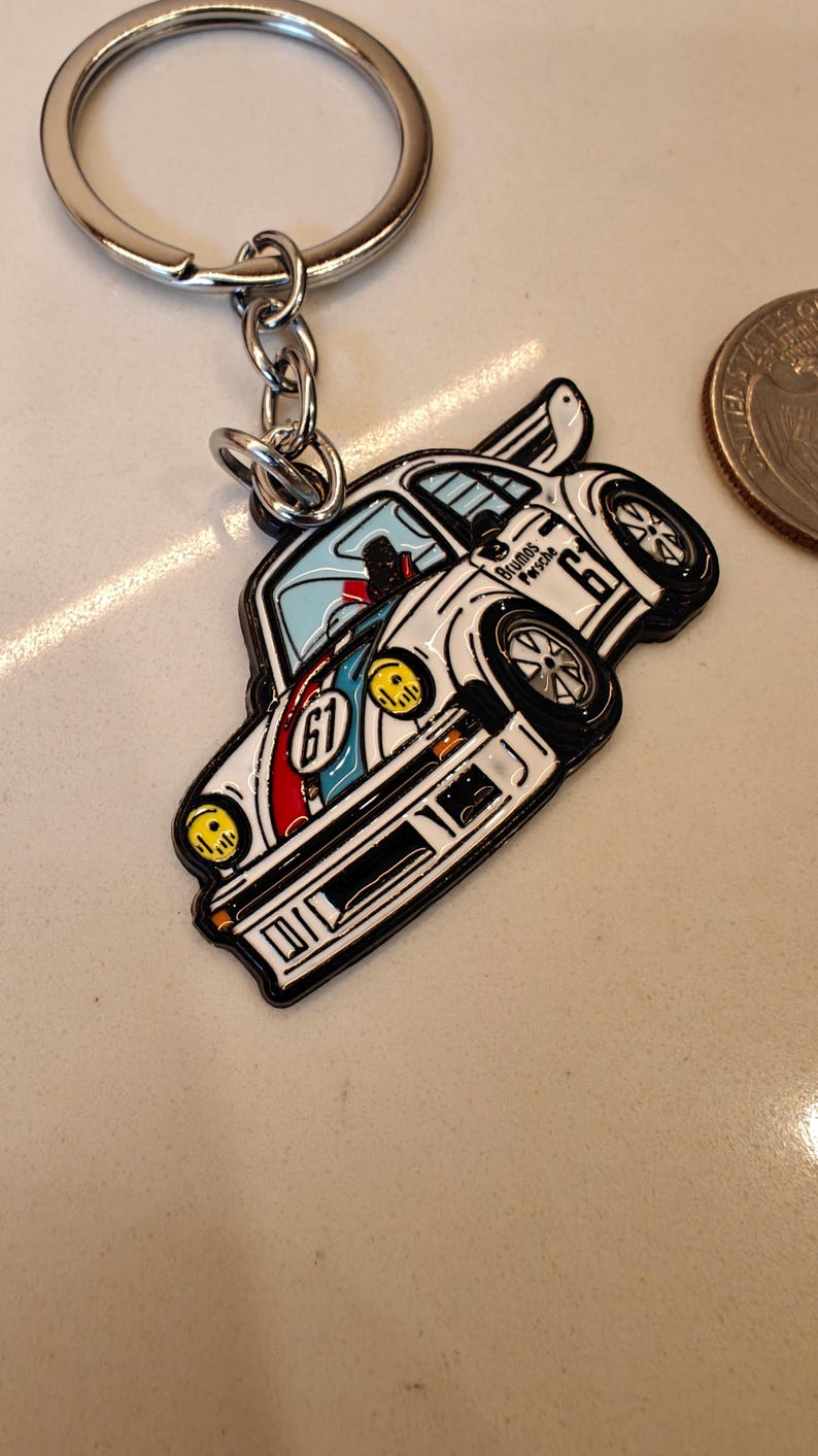 May include: White metal keychain featuring a cartoon depiction of a blue and red race car with the text "Blumos Porsche 67" on the side.