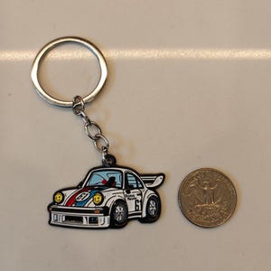 May include: A white and blue car keychain with a red stripe and the text "Brumos Porsche 61" on the side. The keychain is attached to a silver key ring.