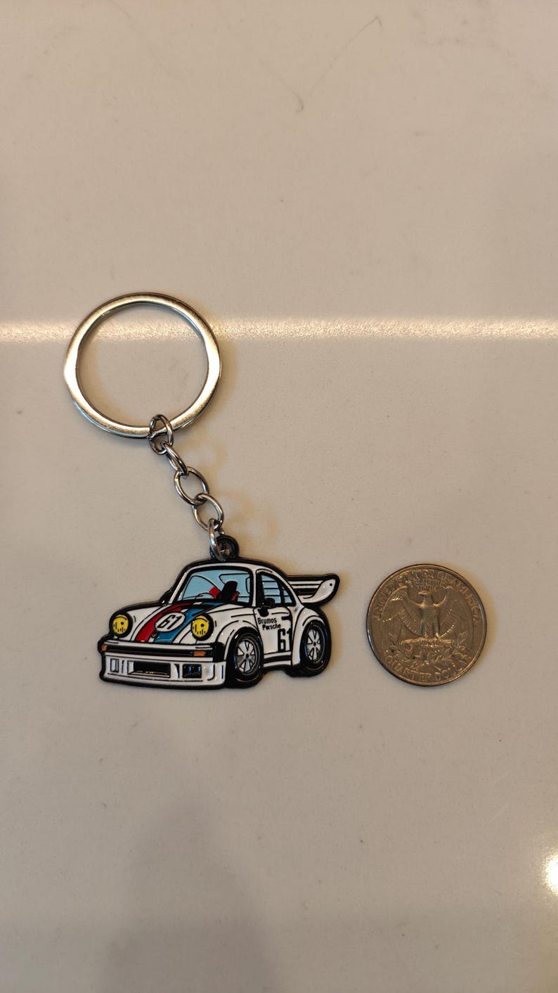 May include: A white and blue car keychain with a red stripe and the text "Brumos Porsche 61" on the side. The keychain is attached to a silver key ring.