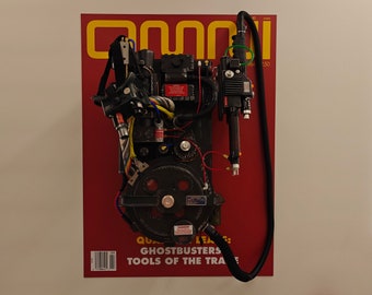May include: A black and silver Ghostbusters proton pack replica, mounted on a red magazine cover with the text "Omni" and "Ghostbusters: Tools of the Trade".