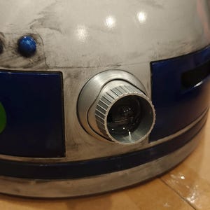 May include: A dome-shaped object with a weathered white and blue finish, featuring a prominent circular lens housing with a ridged silver rim. The surface displays dark smudges and scratches, with small blue circular accents near the top edge.