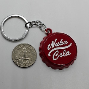 May include: A red Nuka Cola bottle cap keychain with white lettering. The keychain includes a silver key ring and chain. A US quarter is next to the keychain for size comparison.