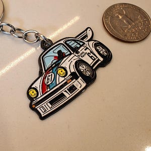 May include: White and blue enamel car keychain with a black outline and the text "Brumos Porsche 67" on the side.