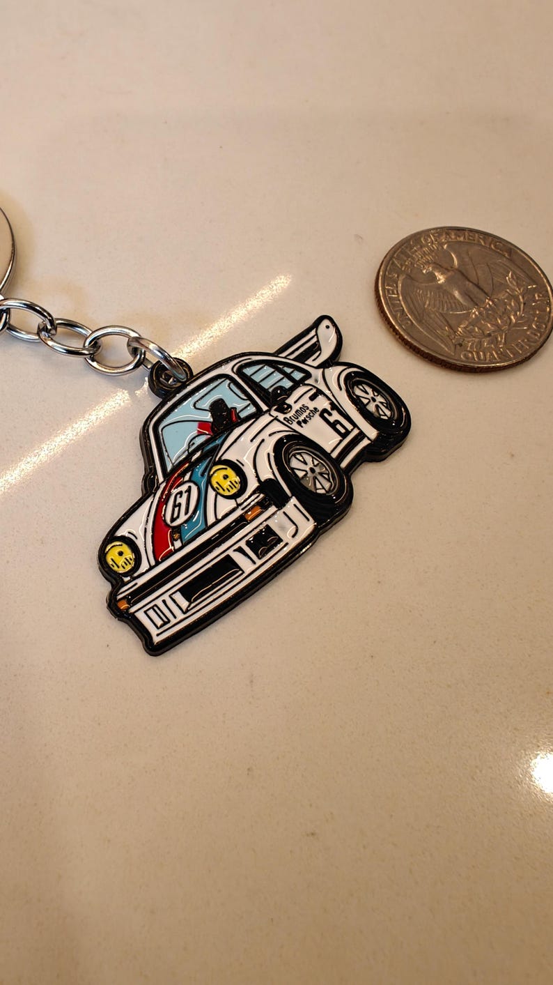 May include: White and blue enamel car keychain with a black outline and the text "Brumos Porsche 67" on the side.