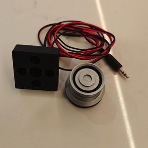 May include: A black rectangular electronic component with five circular indentations, paired with a silver cylindrical sensor featuring a central ring. Both parts are connected by thin red and black wires ending in a standard 3.5mm audio jack plug.