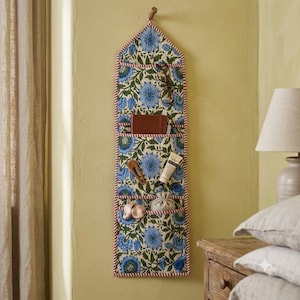 May include: A vertical fabric wall organizer featuring a blue and green floral pattern with red and white striped trim. It has four horizontal pockets containing various small personal items like glasses, a notebook, and a tube of cream.