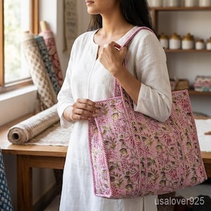 May include: A pink floral tote bag with a quilted design. The bag features a repeating pattern of flowers and leaves in shades of pink, green, and purple. The bag has a rectangular shape with a reinforced top and handles.