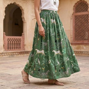 May include: A floor-length green skirt featuring a repeating print of cheetahs, zebras, and tropical palm trees. The skirt has a gathered waistband and a full, flowing silhouette. It is paired with a simple white sleeveless top.