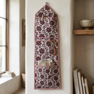 Op de afbeelding: Een verticale stoffen wandorganizer met een donker kastanjebruin bloemenpatroon op een lichtbeige achtergrond. Het heeft drie gestapelde vakken met een rood-witte gestreepte rand, gevuld met pennen, een bril en gevouwen stoffen servetten.