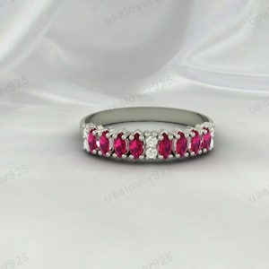 Può includere: Un anello d'argento con una serie di pietre preziose rosse vivaci a taglio marquise alternate a piccole pietre preziose rotonde e chiare. L'anello è posto su uno sfondo bianco strutturato.