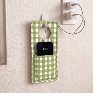 Puede incluir: Un soporte de tela rectangular para cargar el teléfono con un patrón de cuadros vichy en verde claro y blanco. El soporte tiene un recorte circular en la parte superior para colgarlo de un gancho y un bolsillo frontal con un smartphone negro.