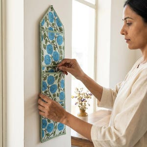 May include: A vertical fabric wall organizer featuring a light green background with a repeating pattern of blue floral motifs and dark green stems. The organizer has three stacked pockets with pointed top edges and green fabric trim borders.