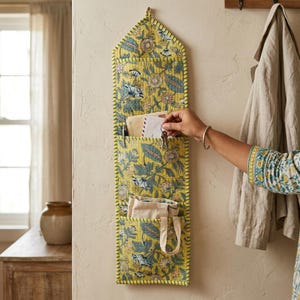 May include: A vertical wall-mounted fabric organizer featuring a yellow background with a blue and green floral pattern. It has three stacked pockets for storage, with the top pocket holding envelopes and the bottom pocket containing a small bag.